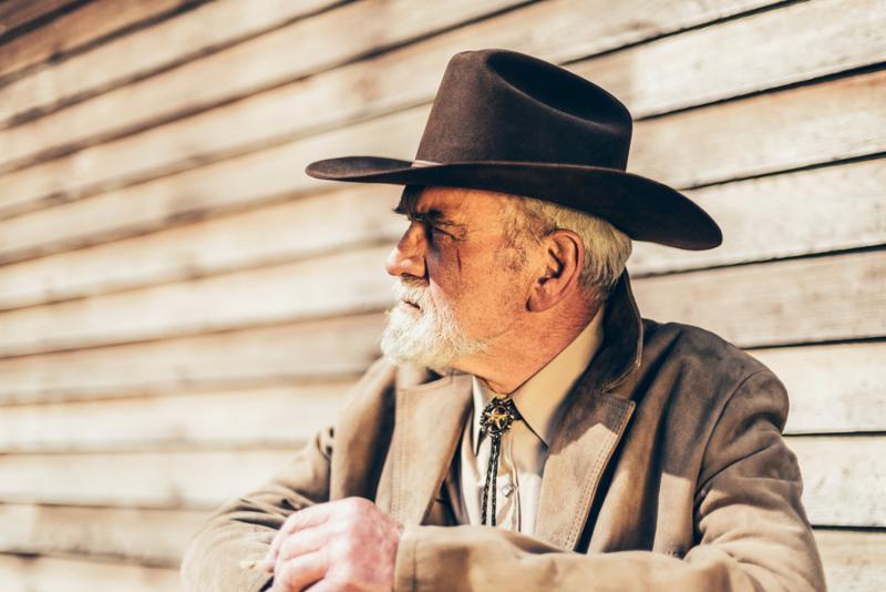 Close up Pensive Old Western Man Wearing Jacket and Cowboy hat Looking to the Left of the Frame While Sitting on the Steps In front of his House.