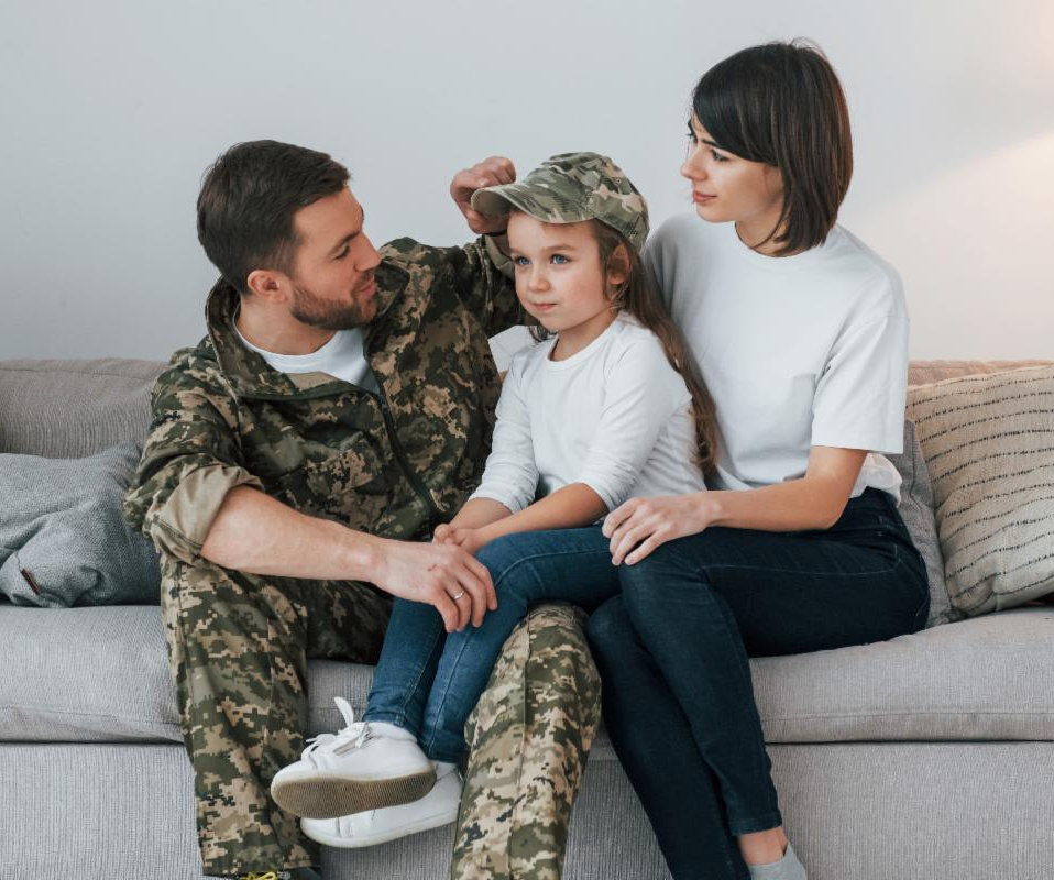 Photo of a soldier in uniform sitting on a grey couch with his wife and young daughter.