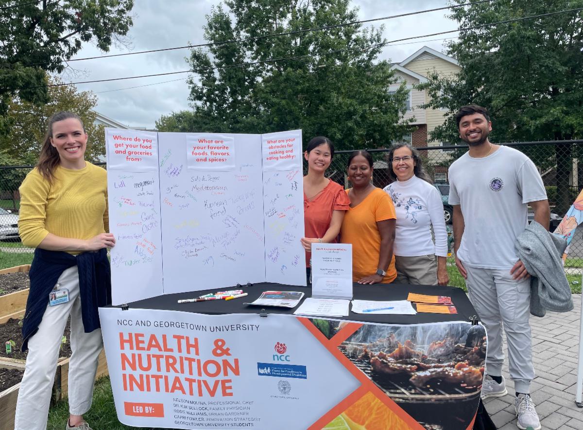 A group of scholars and community members are outside around a table for the Health and Nutrition Initiative
