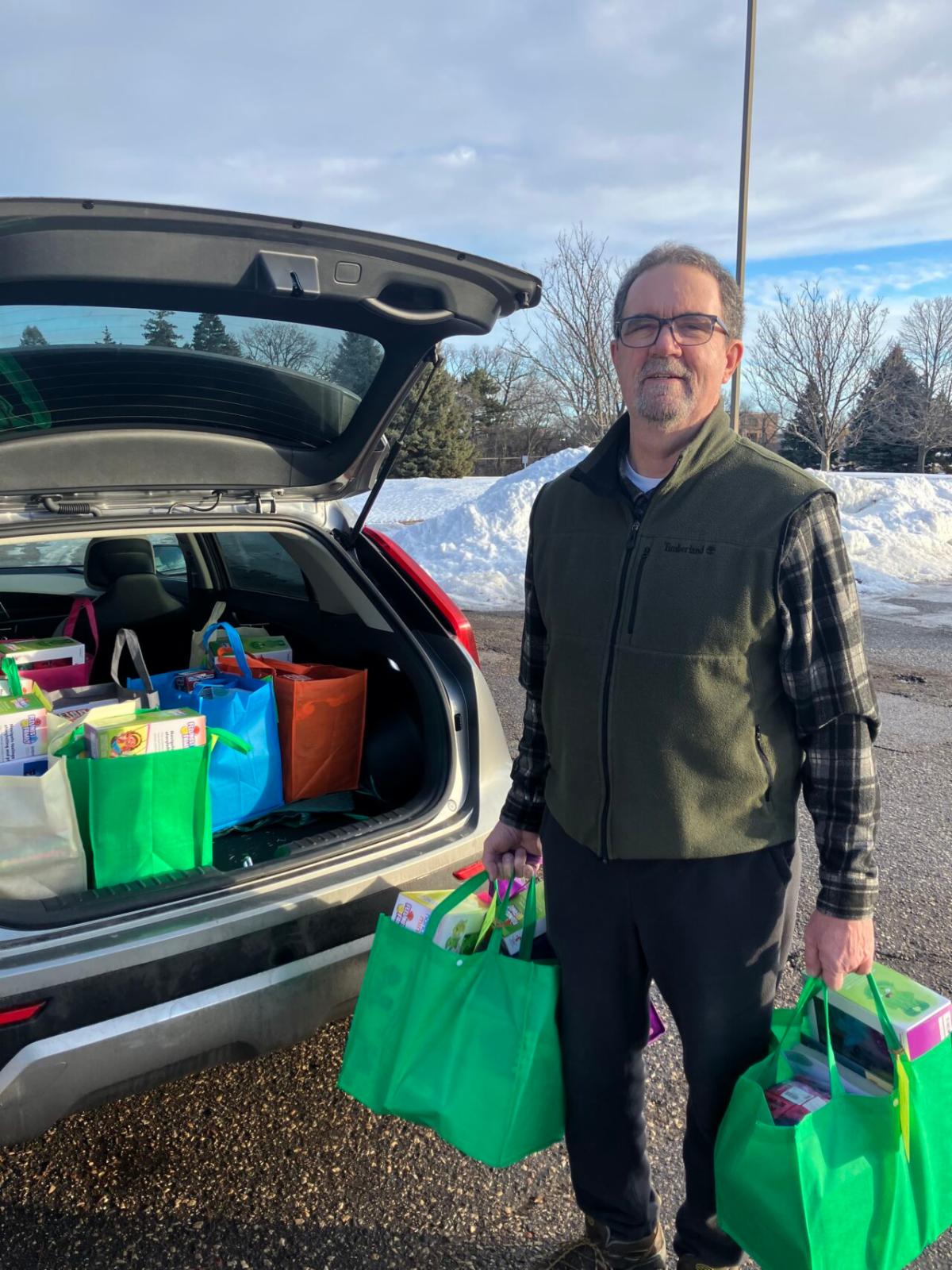 A man stands beside an open car trunk filled with colorful teacher toolkits, holding two bags as he prepares to deliver them to schools.