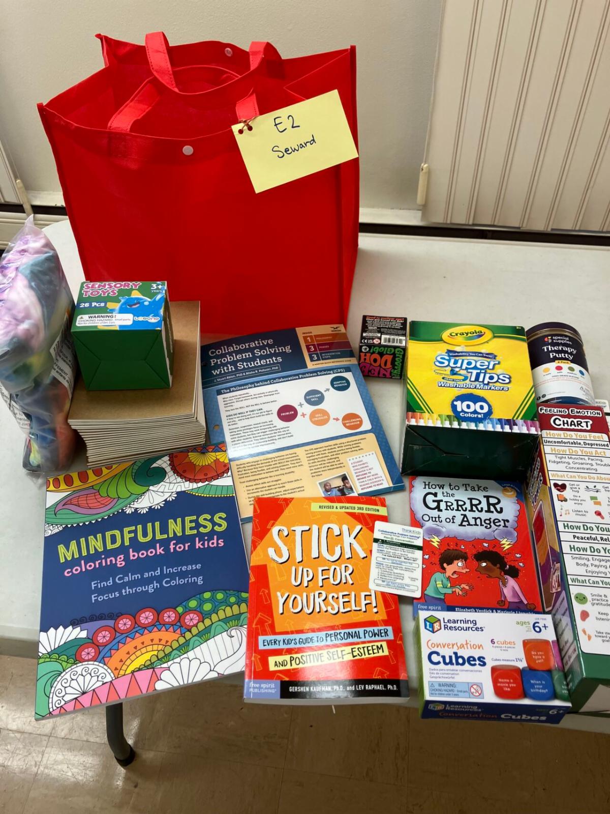 A table displays the contents of a teacher toolkit, including children’s books, journals, sensory toys, markers, therapy putty, conversation cubes, and a red tote labeled for a classroom.