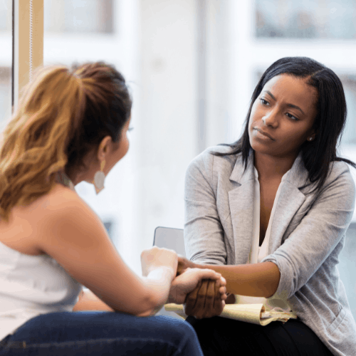 A woman listens attentively while holding another woman’s hand during a calm, supportive conversation.