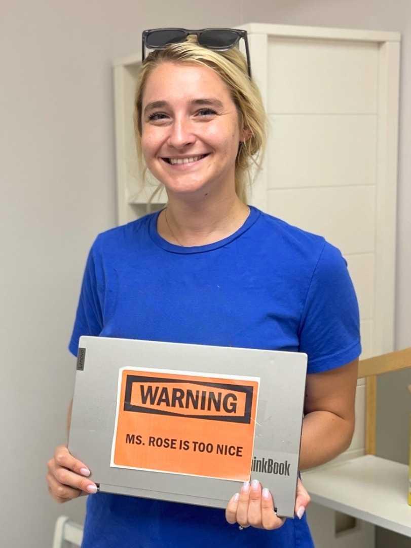Smiling blonde woman in a blue t-shirt holding a laptop with an orange sticker on it that says "WARNING. MS. ROSE IS TOO NICE."