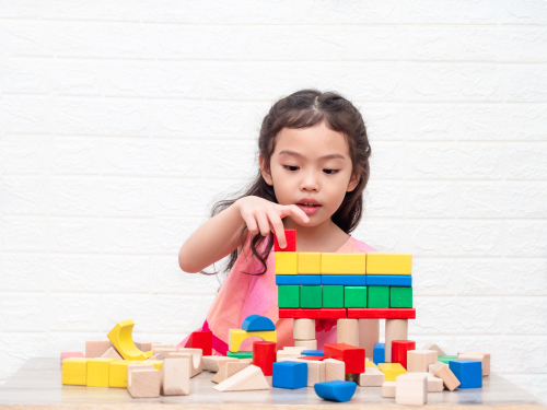 Little cute girl 6 years old playing wooden blocks on table and white bricks wall  background. Learning and education.