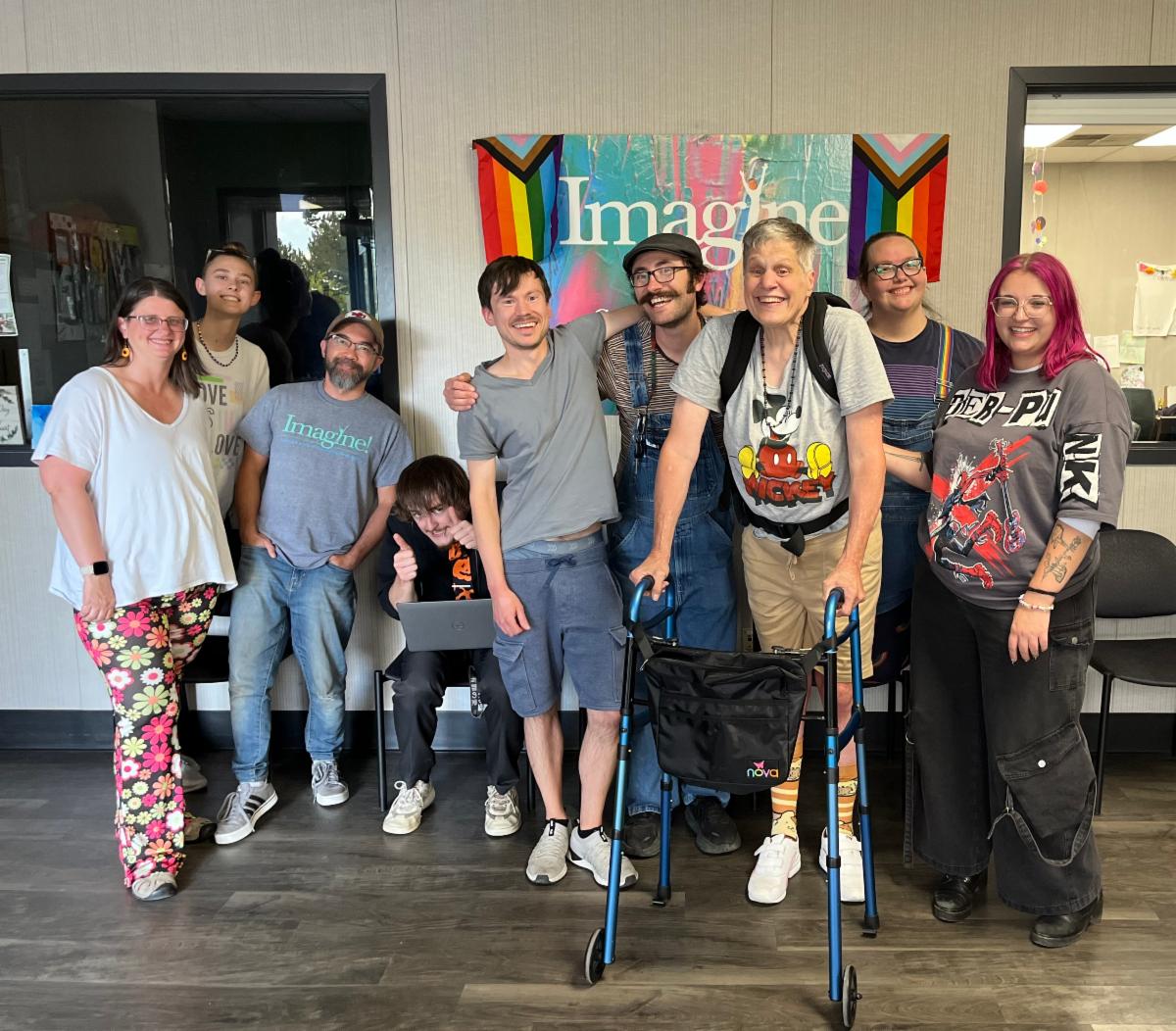 A group of nine people pose together and smile inside of a warm friendly and inclusive space. They stand in front of a colorful Imagine sign with rainbow pride flags on each side. The group includes individuals of different ages and styles. One person uses a walker and one sits with a laptop giving a thumbs up while others stand close together with arms around each other.