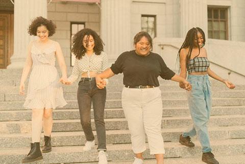 Four females of diverse races are holding hands and smiling and walking down the steps of a building together