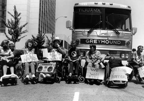 A group of people who are wheelchair users hold protest signs and sit in front of a Greyhound bus