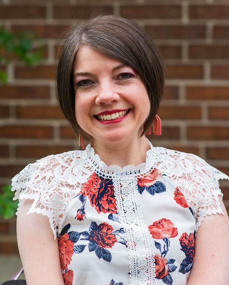 Picture of Carrie from the waist up smiling and wearing a white and peach and blue flowered lacy shirt