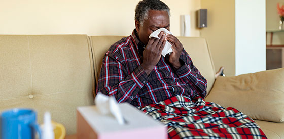 Man in plaid robe sitting on couch blowing his nose.