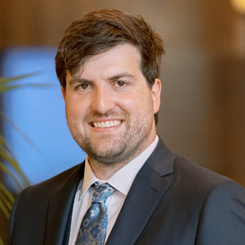 A white man with brown hair smiling with a blue tie and dark grey suite