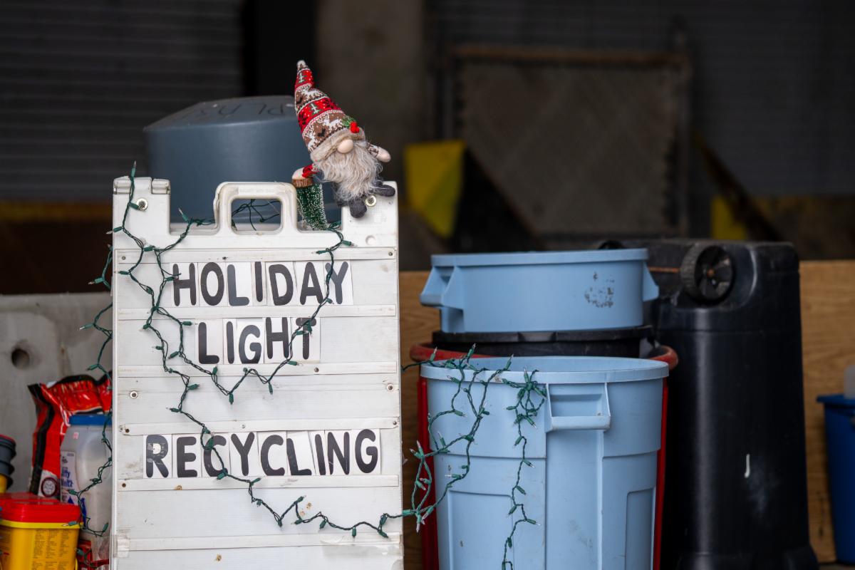 Holiday light recycling sandwhich board sign next to a blue trash bin both are loosely strung with xmas lights and one holiday gnome on top