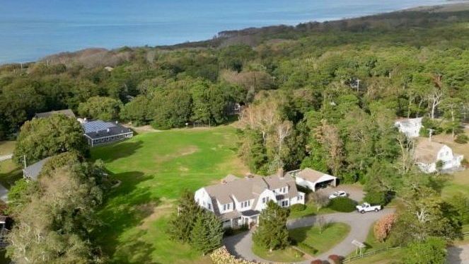 Aerial view of the Bay Property looking northeast. There is a large white house with fields and trees adjacent. The bay is in the background.