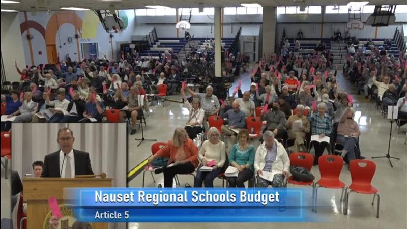 Screenshot of town meeting video showing town meeting body there are about 250 people sitting in chairs many of whom are holding up pink pieces of paper.