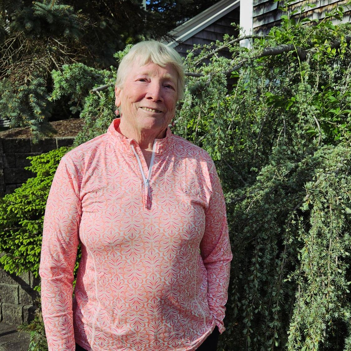 Image is of Anne O'Connell she is wearing a long pink and white patterned shirt and is standing in front of some green ornimental shurbs.