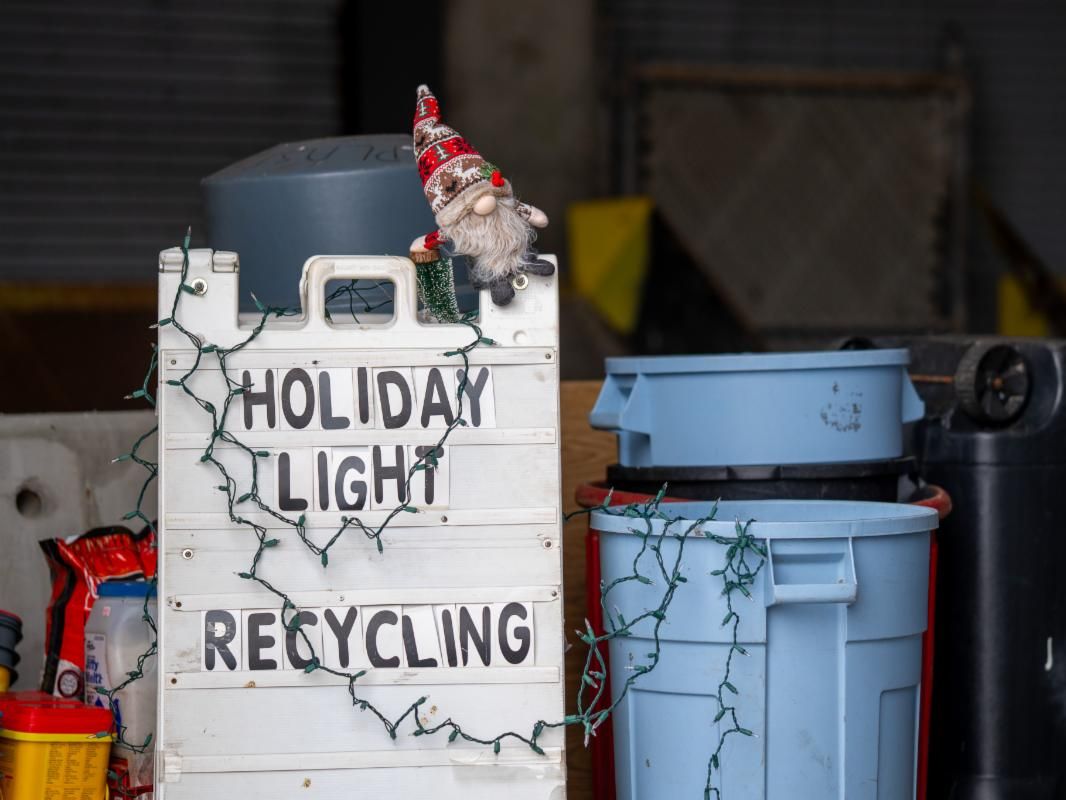 Holiday light recycling sandwhich board sign next to a blue trash bin both are loosely strung with xmas lights and one holiday gnome on top