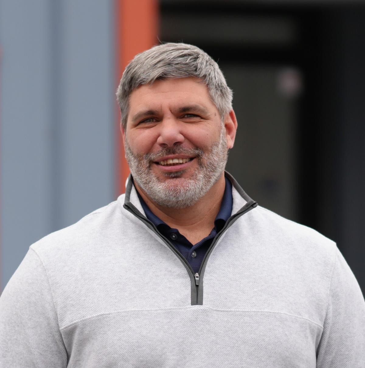 Headshot of Alex Provos standing outside of the DPW bay door wearing a white fleece polo