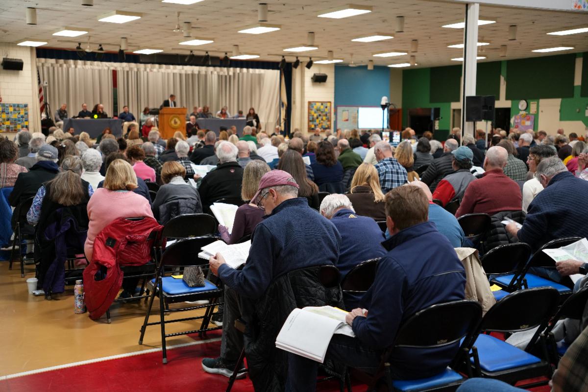 A seated crowd of people in a gym looking toward a stage for town meeting