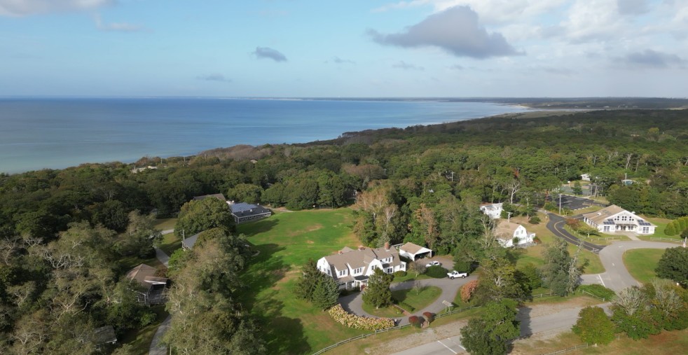Aerial view of the Bay Property looking northeast. There is a large white house with fields and trees adjacent. The bay is in the background.
