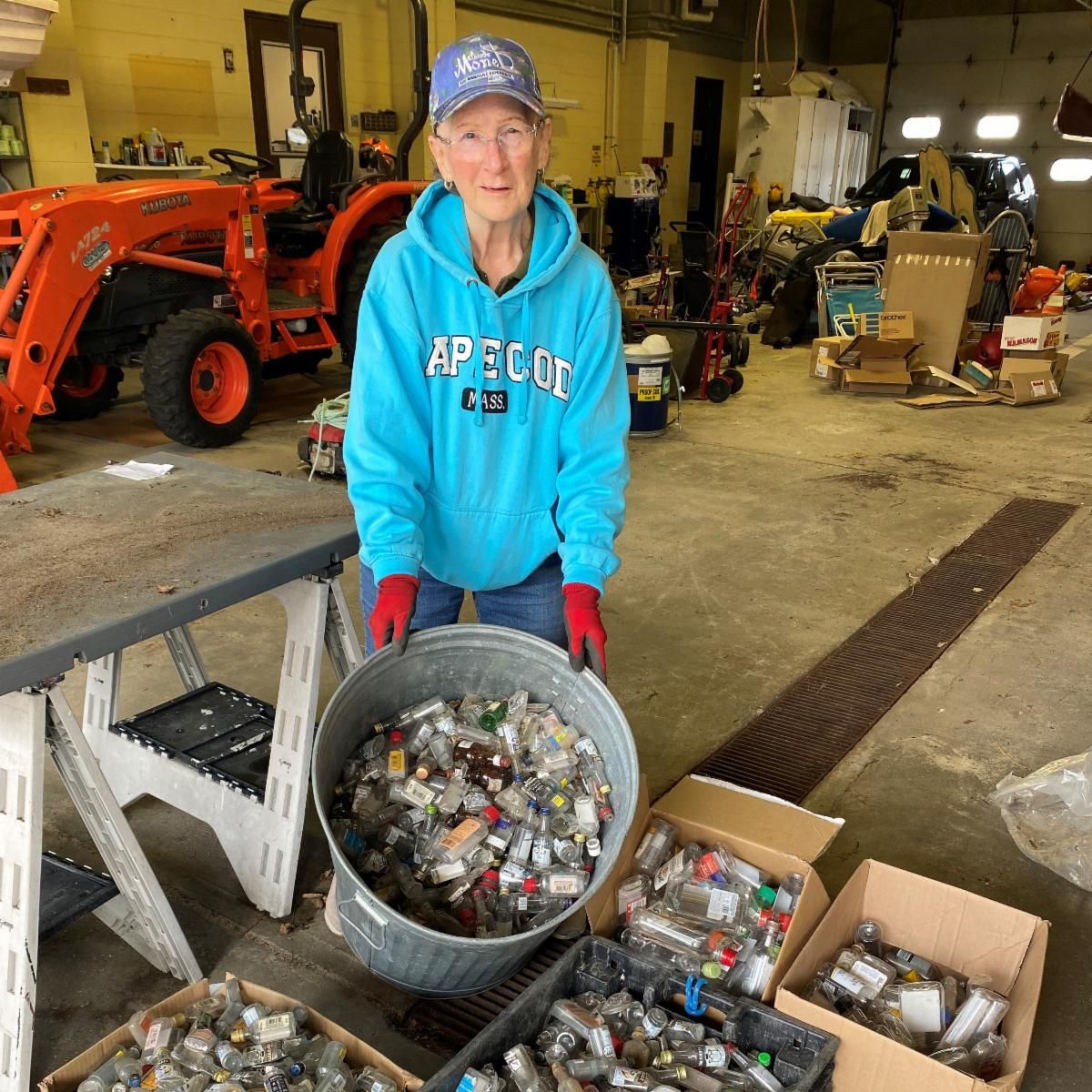 Meg Morris is in a blue sweatshirt and hat tilting a aluminum garbage can will of alcohol nip bottles in front of four other bins also filled with empty nip bottles