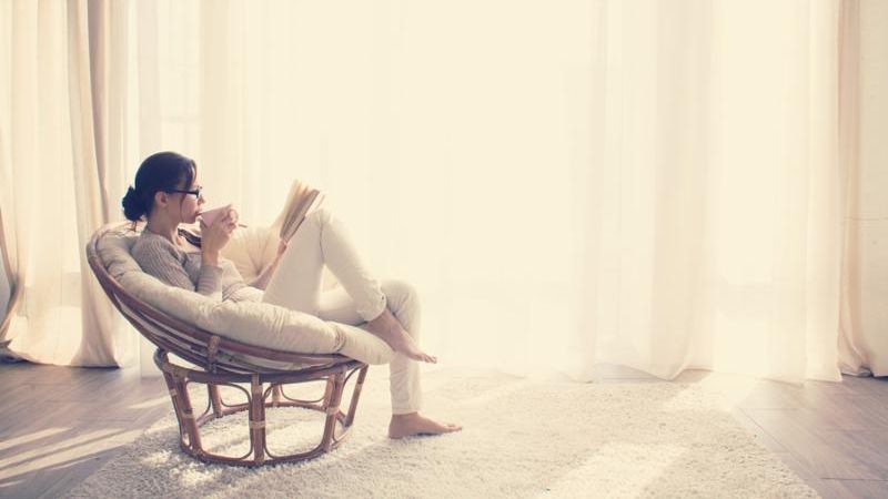 Young woman at home sitting on modern chair in front of window relaxing in her living room reading book and drinking coffee or tea