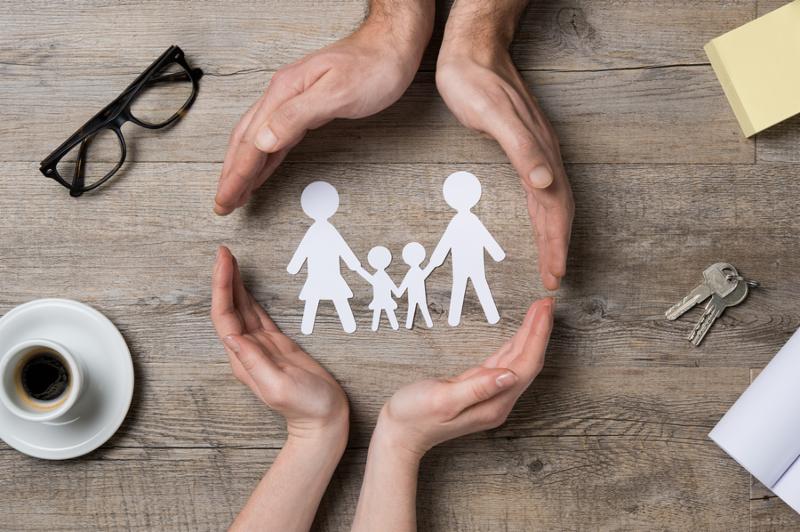 Close up of female and male hands protecting a paper chain family. Top view of two hands form a circle around white paper chain family on wooden table. Family care, insurance and helping hand concept.