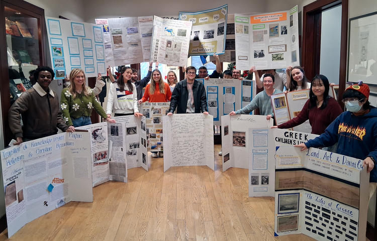 a group of students stand in a building lobby holding posters from their research projects on Hellenic studies topics.