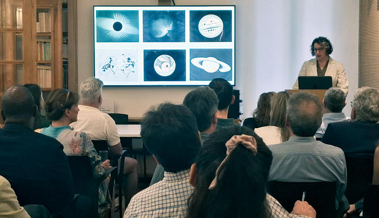 A woman lectures in front of several people next to a screen depicting images of planets including Saturn with rings.