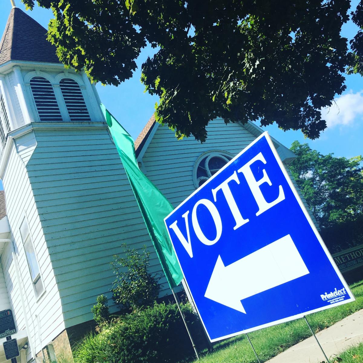 A blue yard sign with a white arrow and white text that reads "VOTE," in the lawn of a white church building