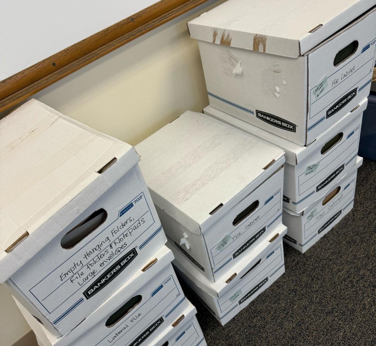 Several stacks of filled file boxes on the floor in front of a wall mounted whiteboard
