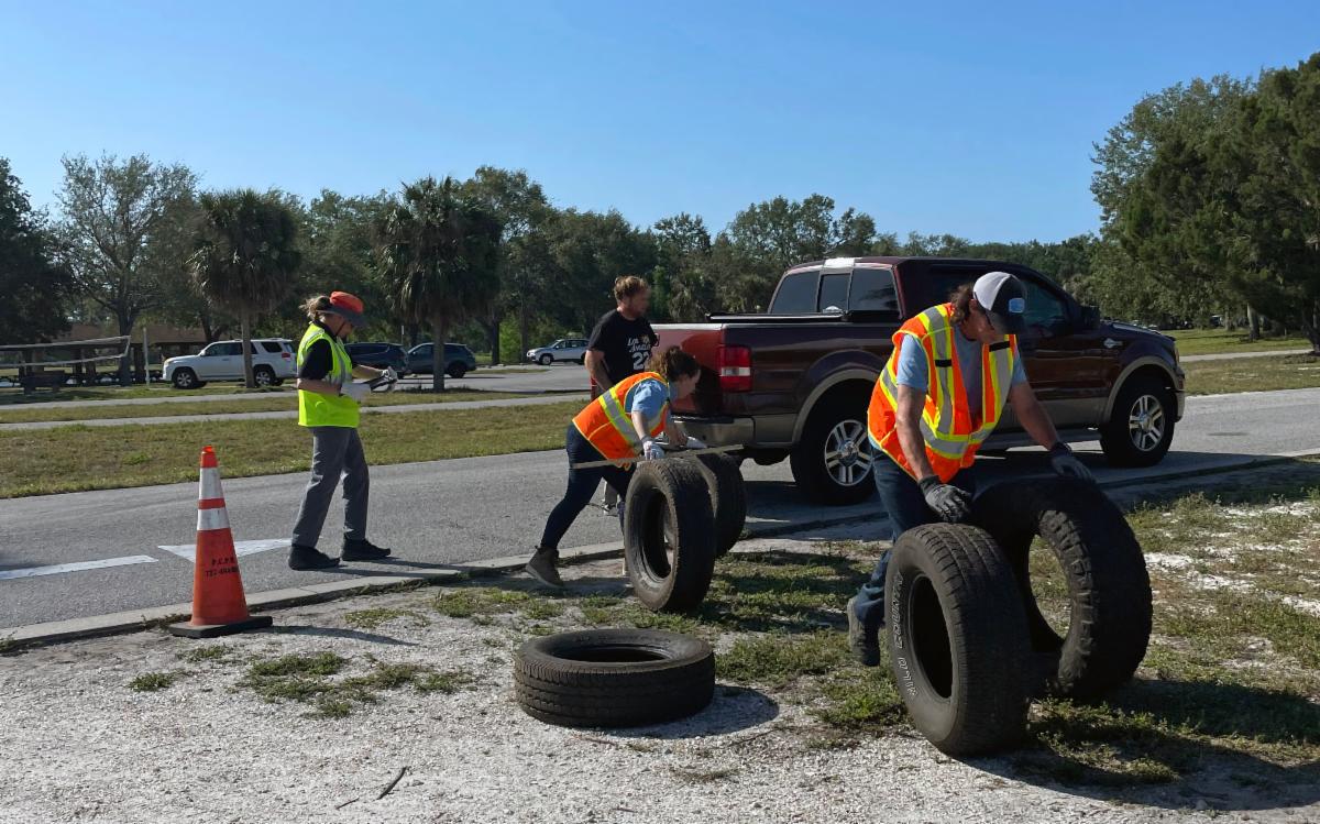 Staff wearing reflective safety vests collect and move discarded tires at a Pinellas County free tire disposal event. Two people roll large tires away from a pickup truck, while another looks at a clipboard.