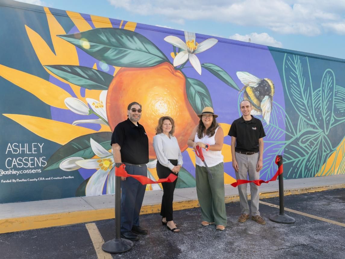 Lealman CRA team cutting ribbon in front a mural of an orange with white flowers around it.