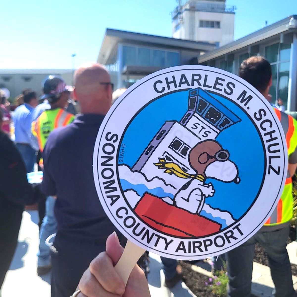 Photo of a Sonoma County Airport logo held by a hand in front of people standing in front of the airport terminal - Foto del logotipo del aeropuerto del condado de Sonoma sostenido con una mano frente a personas paradas frente a la terminal del aeropuerto.