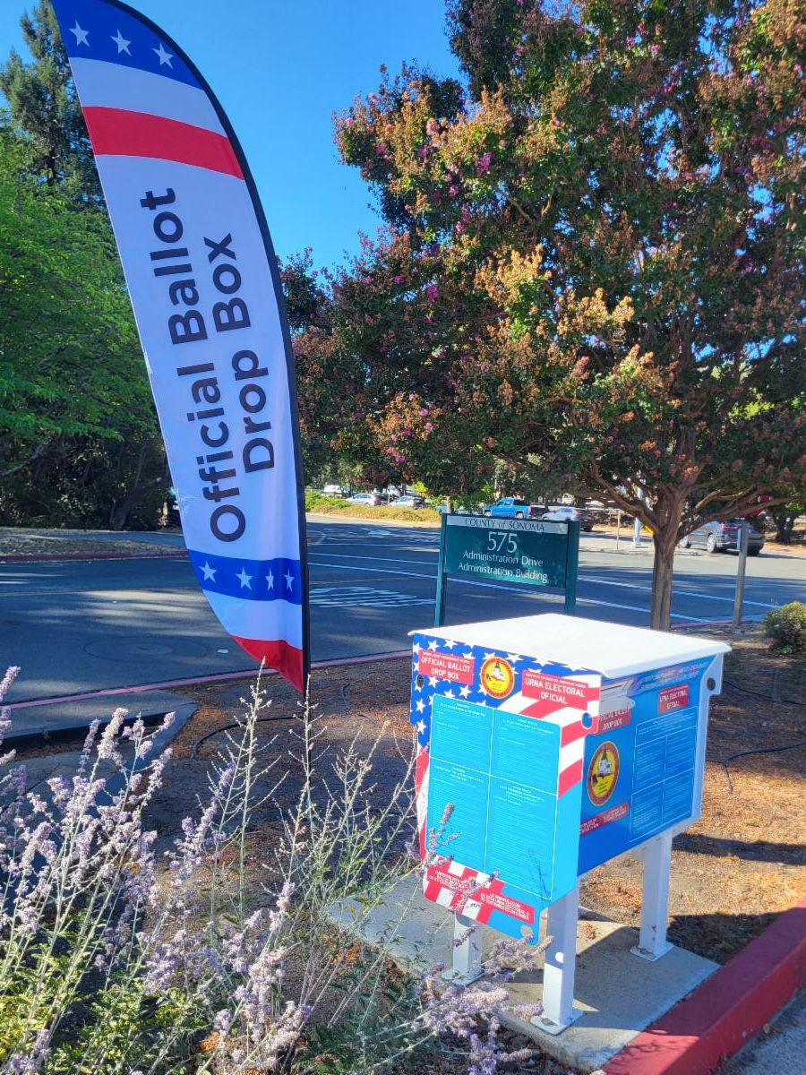 Photo of the Registrar of Voters ballot drop box with vegetation on a drive thru curb at 575 Administration Drive with a banner flag indicating official ballot drop box