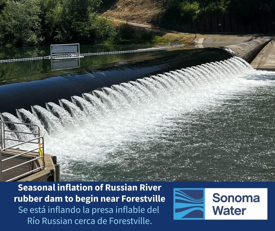 mage of the Russian River rubber dam in Forestville during seasonal inflation. Water is visibly cascading over the inflated black rubber dam under clear skies, with trees and a sloped hillside in the background. A metal railing is seen in the foreground. A sign at the bottom reads: “Seasonal inflation of Russian River rubber dam to begin near Forestville / Se está inflando la presa inflable del Río Russian cerca de Forestville.” The Sonoma Water logo appears in the lower right corner.