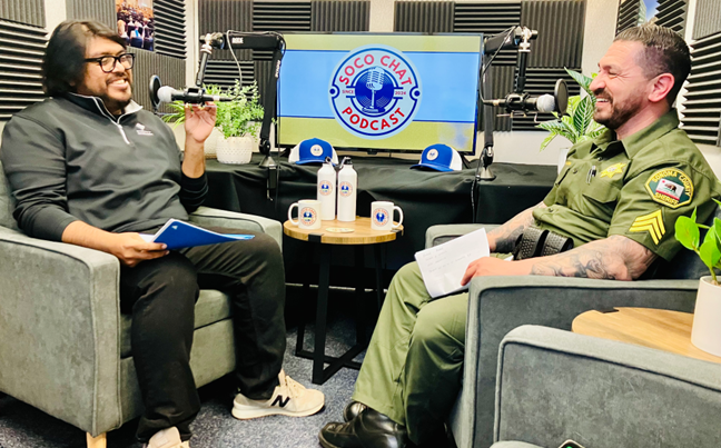Dos hombres incluido el sargento del sheriff sentados en el estudio del podcast SoCo Chat conversando sobre un episodio con el logotipo del podcast en una pantalla al fondo