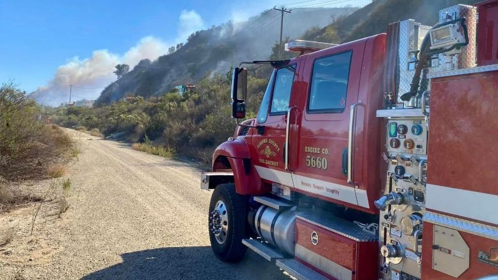 Photo of a firetruck driving on a dirt road approaching a fire on the hillside