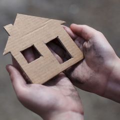 Photo of dirty hands holding a small house cardboard cutout. Foto de unas manos sucias sujetando un recorte de cartón de una casita.