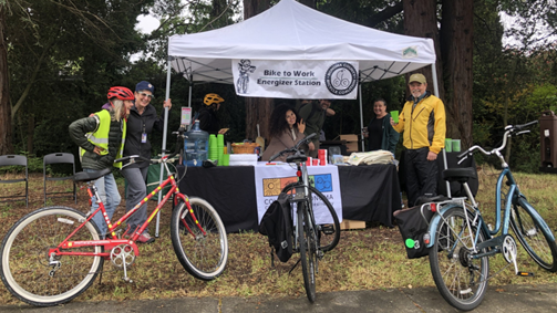 Photo of three bikes and bikers in front of a display table drinking water for bike to work day ~ Foto de tres bicicletas y ciclistas delante de una mesa de exposición bebiendo agua para el día de la bicicleta al trabajo