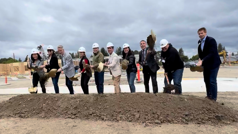 Officials using shovels to dig a pile of dirt at a construction site beneath gray skies. Funcionarios cavan con palas un montón de tierra en una obra bajo un cielo gris.