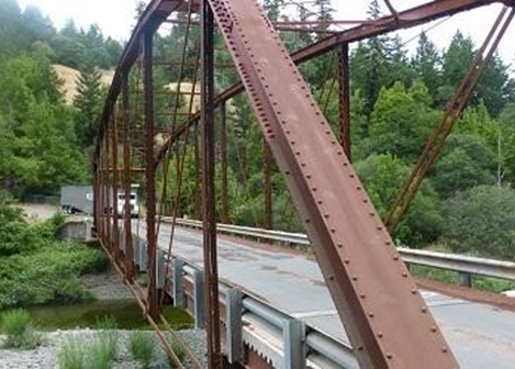Photo of a brown steel bridge over a small river with a big rig about to cross and tree covered mountains in the background