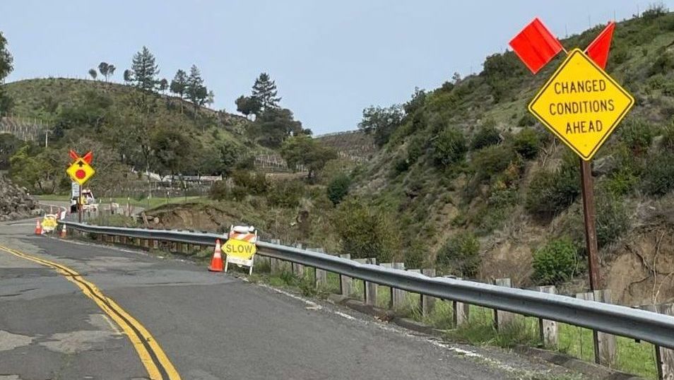 Photo of a hillside road with potholes and a guardrail and road work signs to slow down due to changed conditions ahead