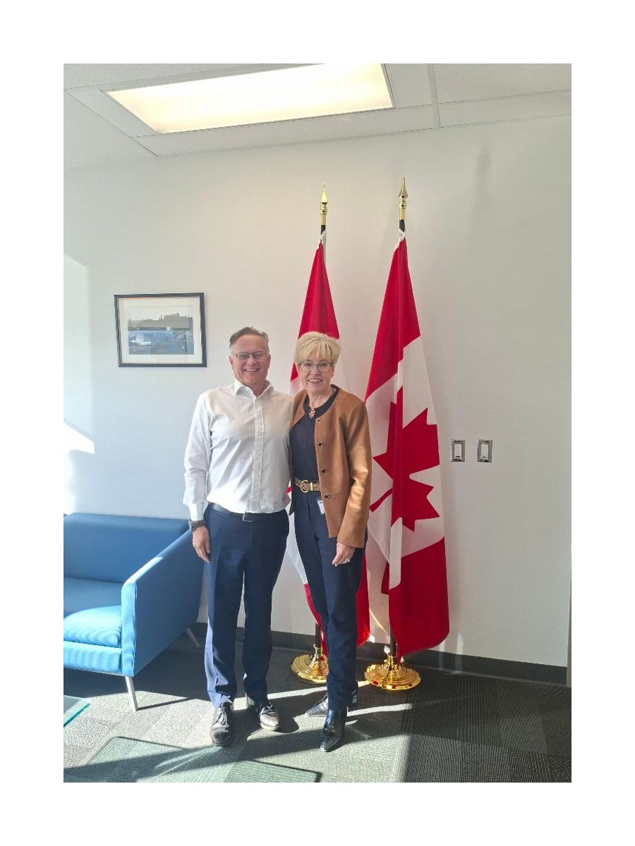 Krista Carr, CEO of Inclusion Canada, stands alongside MP Wayne Long in an office setting, with two Canadian flags displayed behind them. Both are smiling and facing the camera. A blue armchair, framed photograph, and office furnishings are visible in the background.