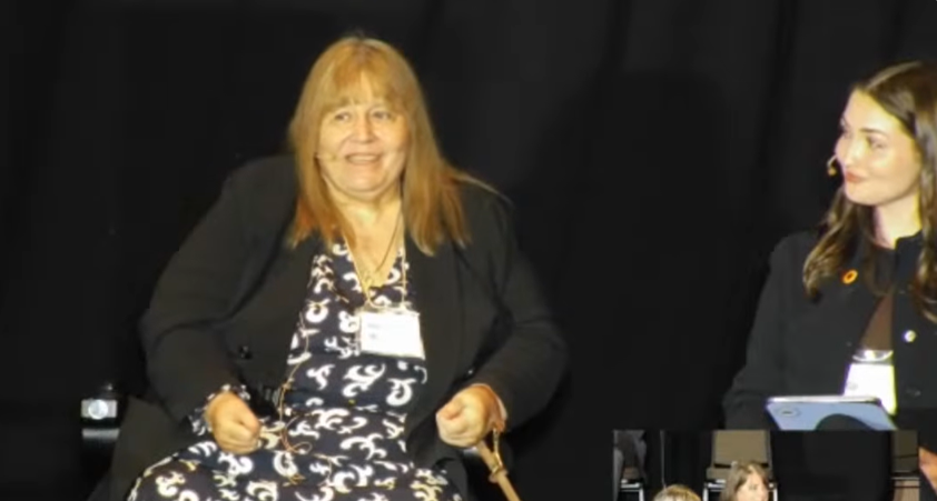 Two women sit on a stage in front of a black backdrop. The woman on the left is seated in a wheelchair and is wearing a black blazer over a patterned dress. The woman on the right is seated beside her and appears to be holding a notepad or papers. Both are facing toward the audience.