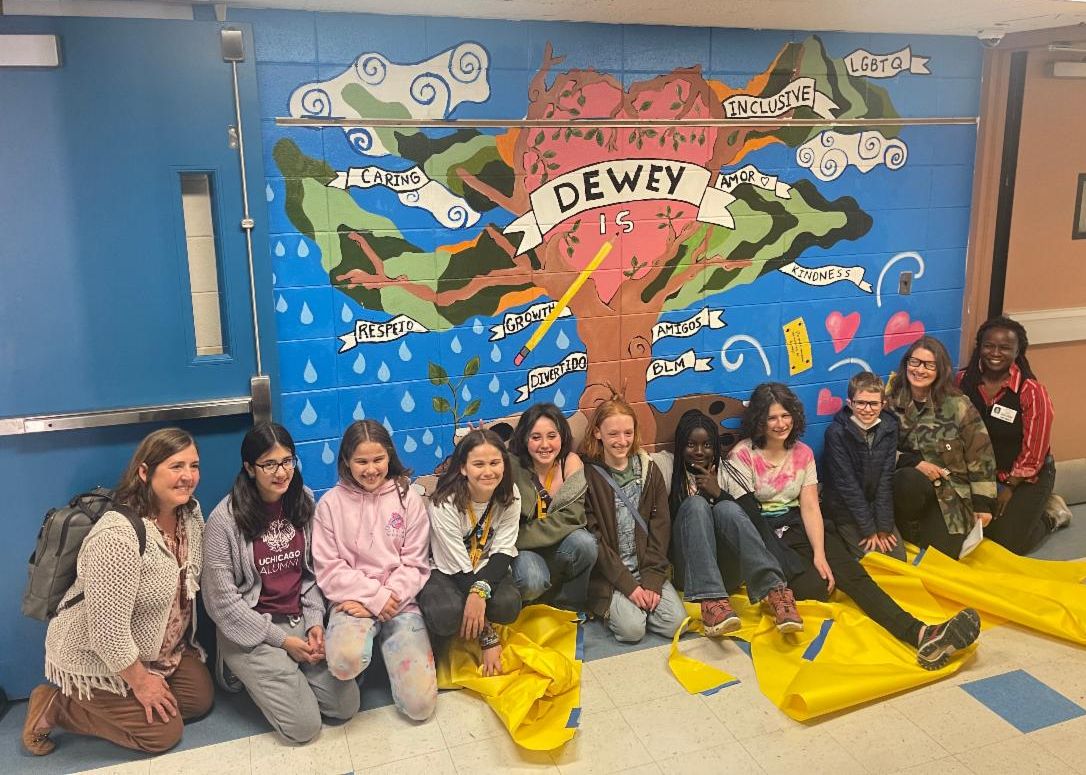 The new Dewey Elementary mural features a tree with a heart at its center, surrounded by branches and words describing the school. Eight students and three adults who assisted with the mural sit in front of it. 