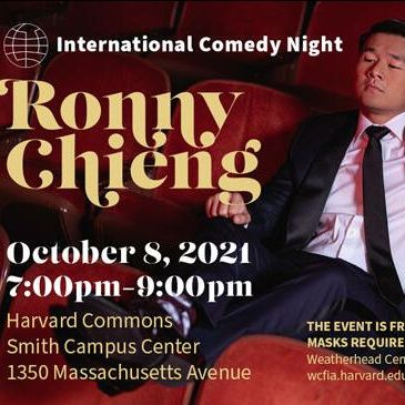 promotional photo of ronny chieng in black suit sitting in red theatre seat with arm draped over the chair next to him