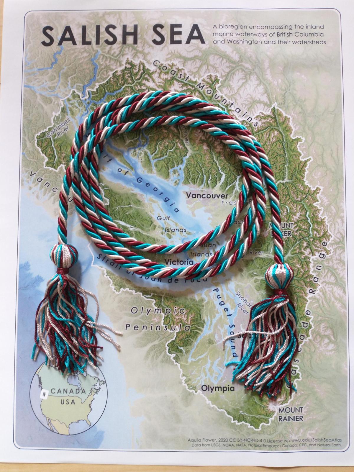 A coil of woven cords that are teal maroon and silver in color at each end is a knot with tassels hanging from them sitting on top of a map of the Salish Sea Region