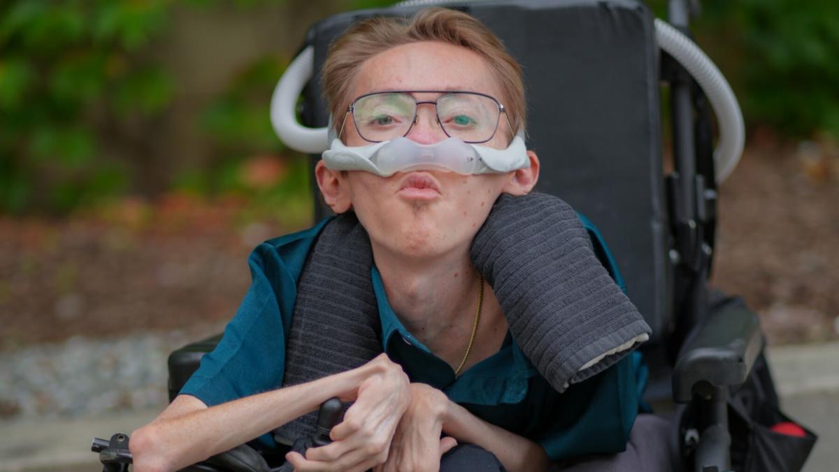 A young white man with muscular dystrophy sitting in a wheelchair with a breathing tube.