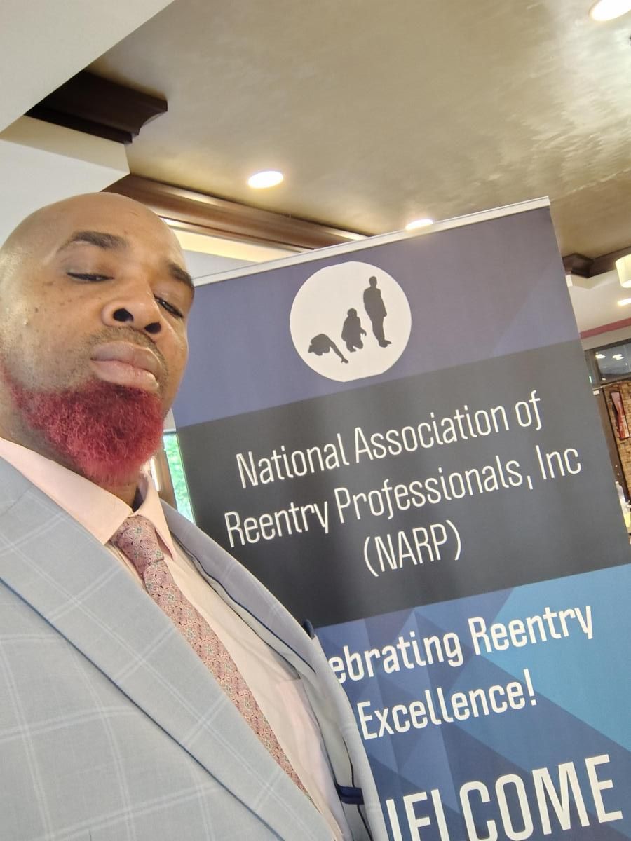 A black man with a red beard stands in front of a poster reading National Association of Reentry Professionals, Inc NARP.