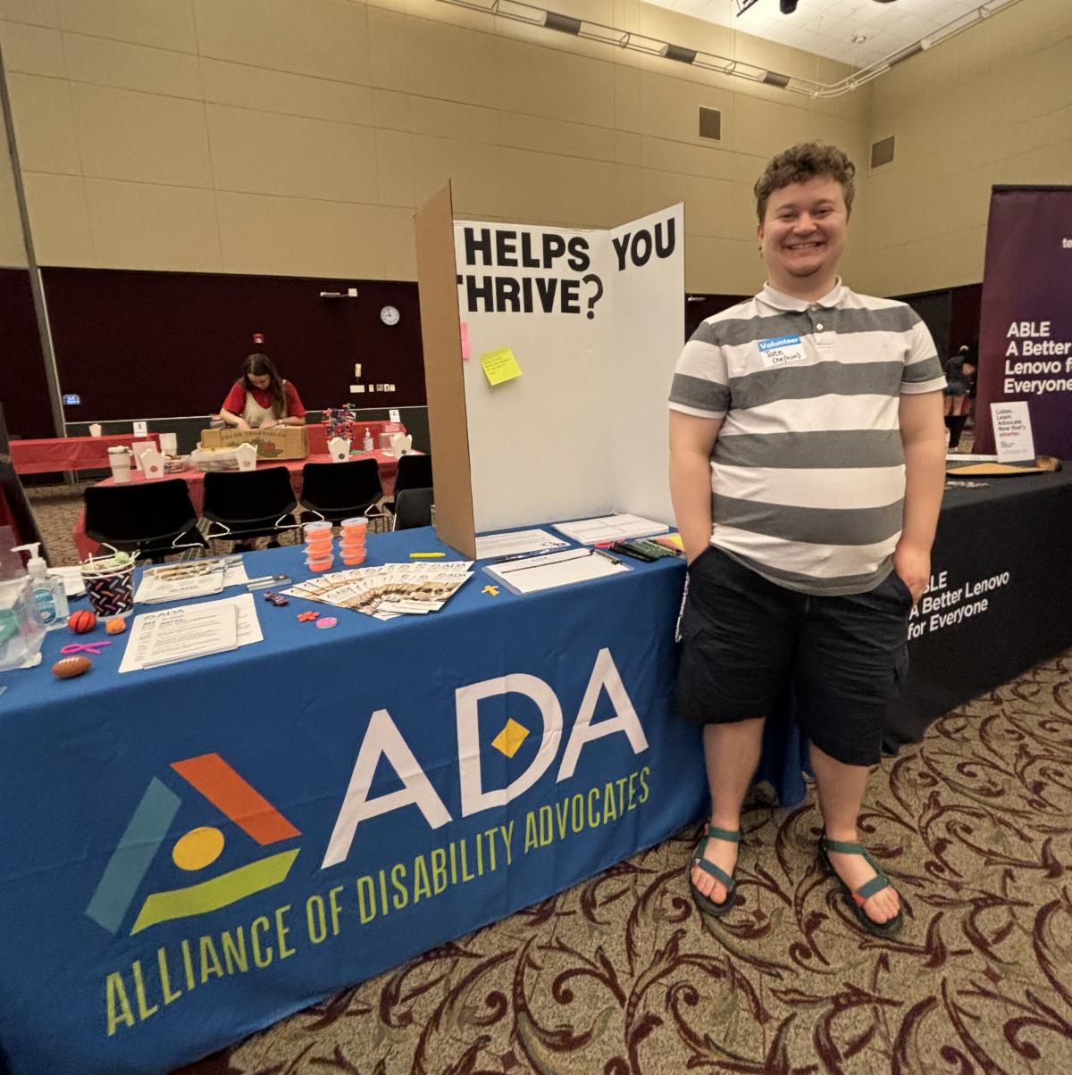 Resource table with a variety of handouts and activities. On the table in a poster board with the text HELPS YOU THRIVE. Smiling next to the table is a young white man.