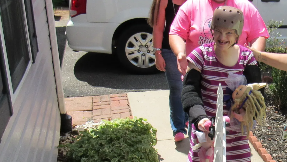 A young white disabled woman wearing a helmet and holding several toys walks down a path into a house.